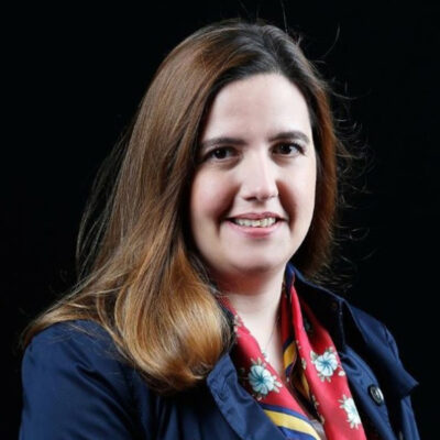 Mulher branca de cabelos castanhos soltos, usando blusa azul e lenço vermelho no pescoço. Atrás, fundo preto.