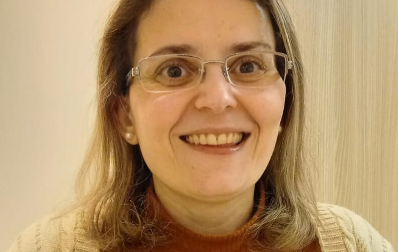 Mulher branca de cabelos loiros soltos sorrindo e usando blusa laranja e casaco creme.