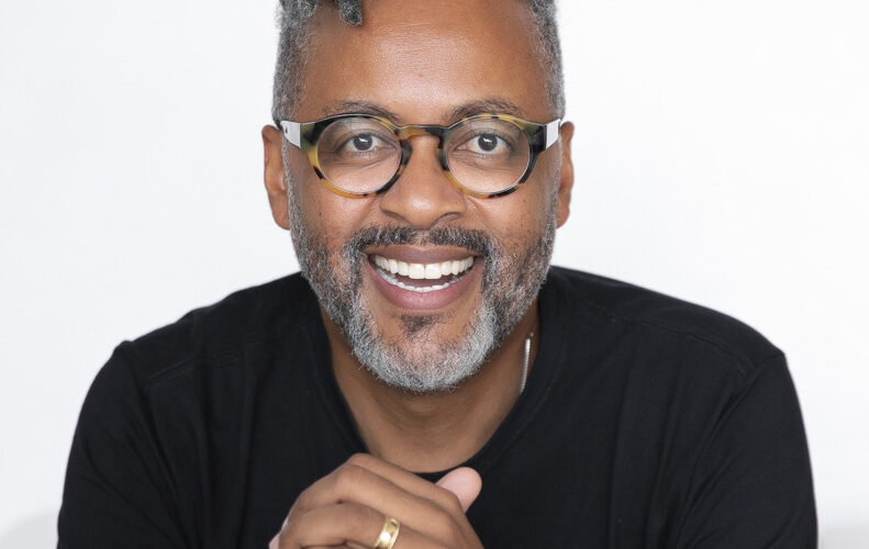 Homem negro usando óculos e blusa preta, sorrindo sobre fundo branco.