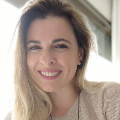 Mulher branca de cabelos loiros soltos, sorrindo e usando blusa creme.