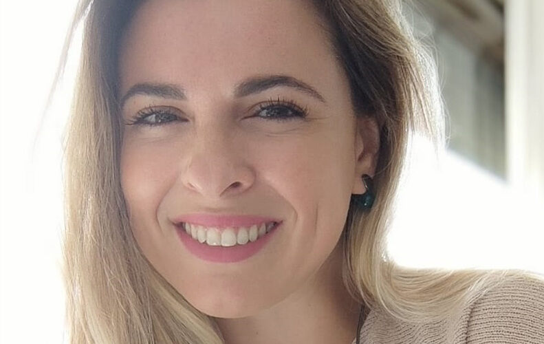 Mulher branca de cabelos loiros soltos, sorrindo e usando blusa creme.