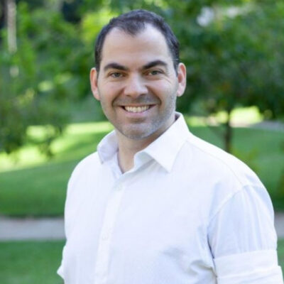 Homem branco sorrindo e usando camisa branca. Atrás, fundo de locar arborizado.