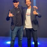 Alex Duarte e Henri Zylberstajn de pé, abraçando-se e mostrando os polegares para cima (positivo).