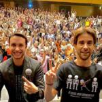 Alex Duarte e Henri Zylberstajn sorrindo. Atrás, público com braços levantados.