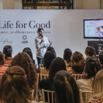 Público de costas assistindo orador Henri Zylberstajn, vestindo camisa branca e na frente de painel branco com texto "Life for Good".