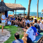 Grupo de pessoas sentadas em meia lua com Henri Zylberstajn no meio em lugar aberto e paisagem de praia no plano de fundo.