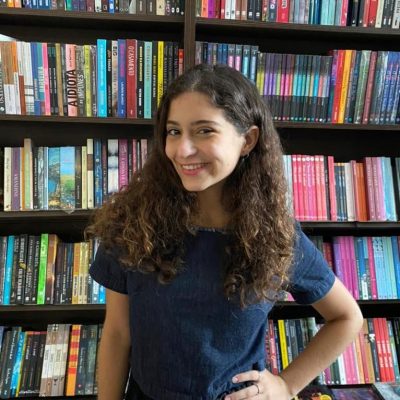 Mulher branca de cabelos longos soltos, sorrindo e usando blusa azul escura. Atrás, estante de livros.
